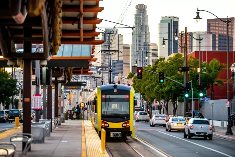 Yellow and black Metro light rail train pulling up to outdoor platform with Los Angeles skyline in background.